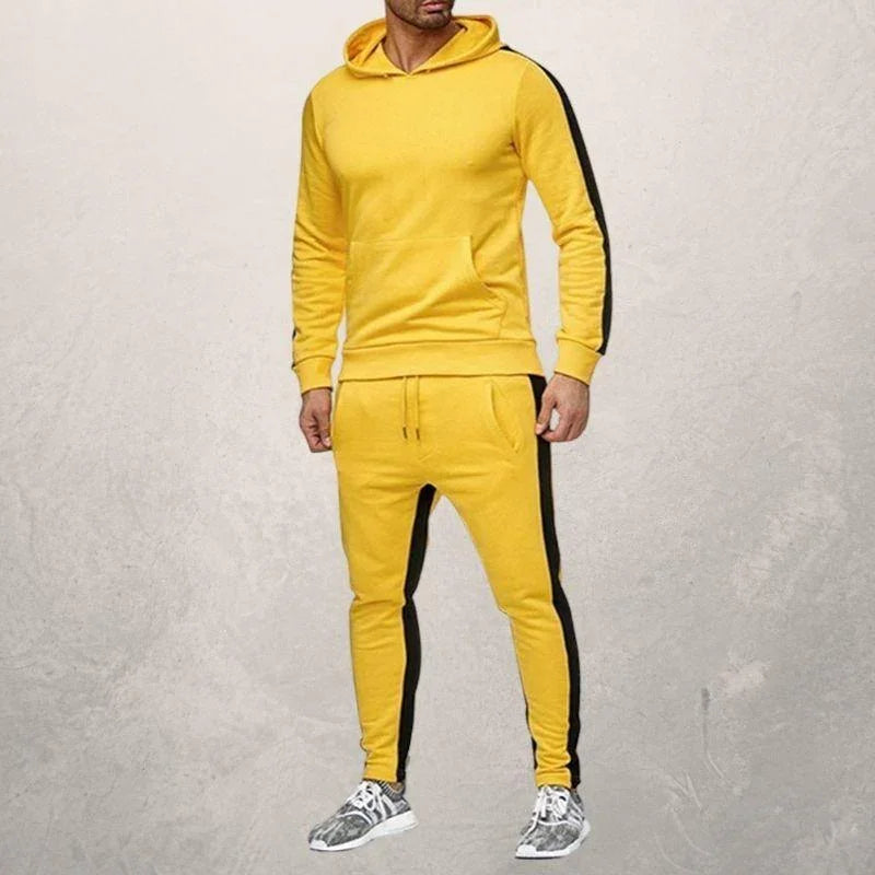 Man wearing yellow fitness hoodie and joggers with black side stripes, standing against gray wall