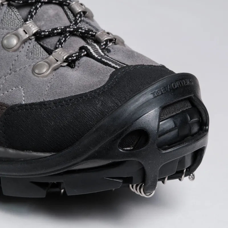 Close-up of a gray and black hiking boot with metal crampons attached to the sole, photographed on a light gray studio background. The brand name TORQOR is embossed on the black toe bumper.