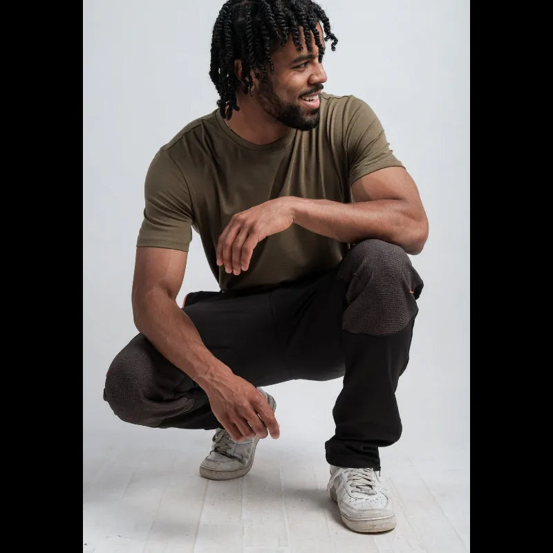 A man with dreadlocks crouching in a studio while wearing an olive green crew-neck t-shirt, dark brown sweatpants with contrast knee patches, and white sneakers.