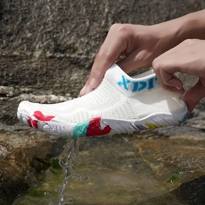 A white slip-on mesh shoe with colorful red, teal, and yellow graphic accents, held by hands over a stream of water with a rocky background.