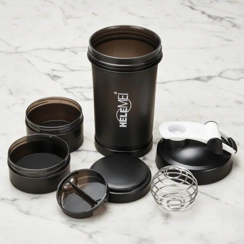 A black metal shaker bottle with its various accessories arranged on a white marble surface. The accessories include two small round storage jars, a black screw-on lid, a clear wire strainer, and a white-and-black shaker spoon.