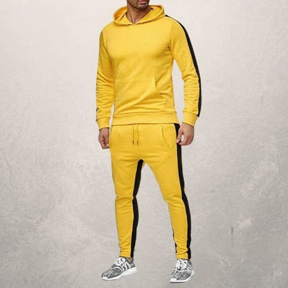 Man wearing yellow fitness hoodie and joggers with black side stripes, standing against gray wall