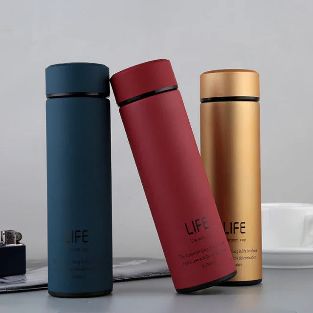 Three sleek LIFE vacuum insulated bottles in blue, red, and gold on a gray surface with a white cup and lighter nearby