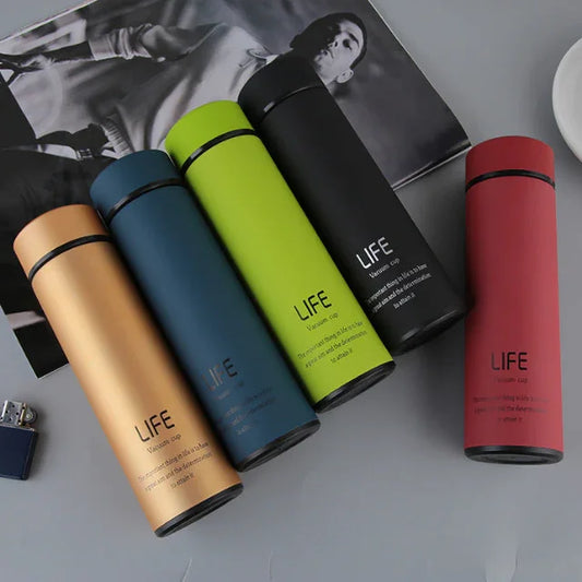 Set of five colorful LIFE vacuum insulated water bottles in gold, blue, green, black, and red on a gray surface with a magazine and lighter