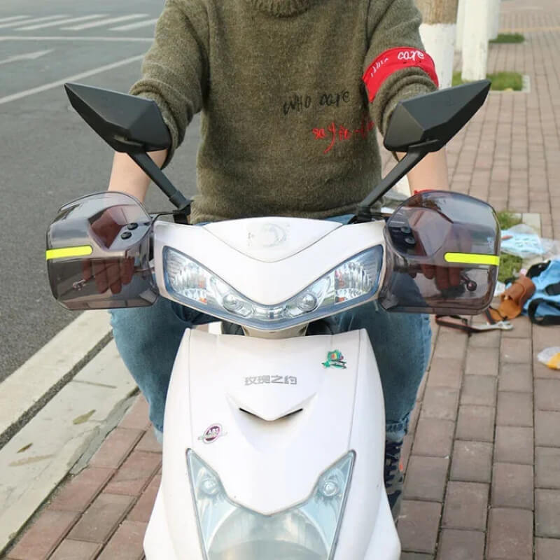 Person riding a white scooter with protective hand guards on the handlebars, city street background