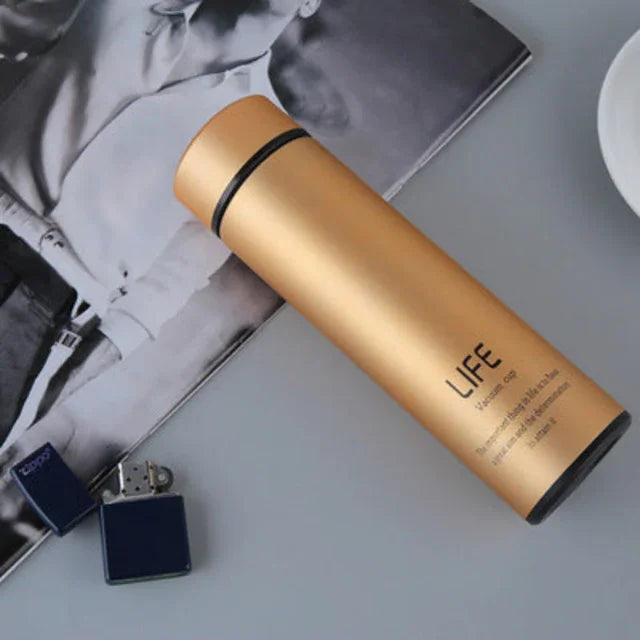 Gold insulated water bottle labeled LIFE on a gray surface with a black and white magazine and navy blue lighter