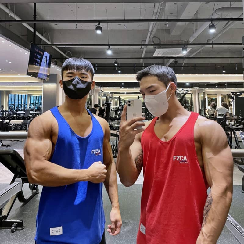 Two muscular individuals in blue and red tank tops taking a selfie in a gym.