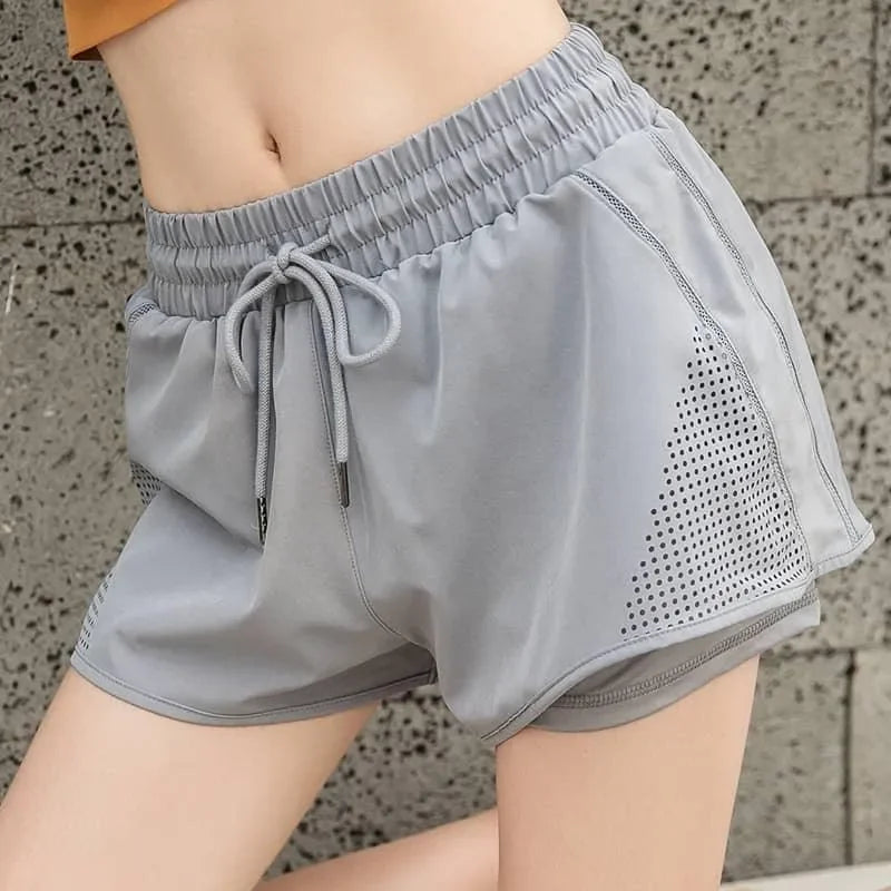 Light gray athletic shorts with an elastic drawstring waist and perforated side panels, shown on a model against a textured gray wall.