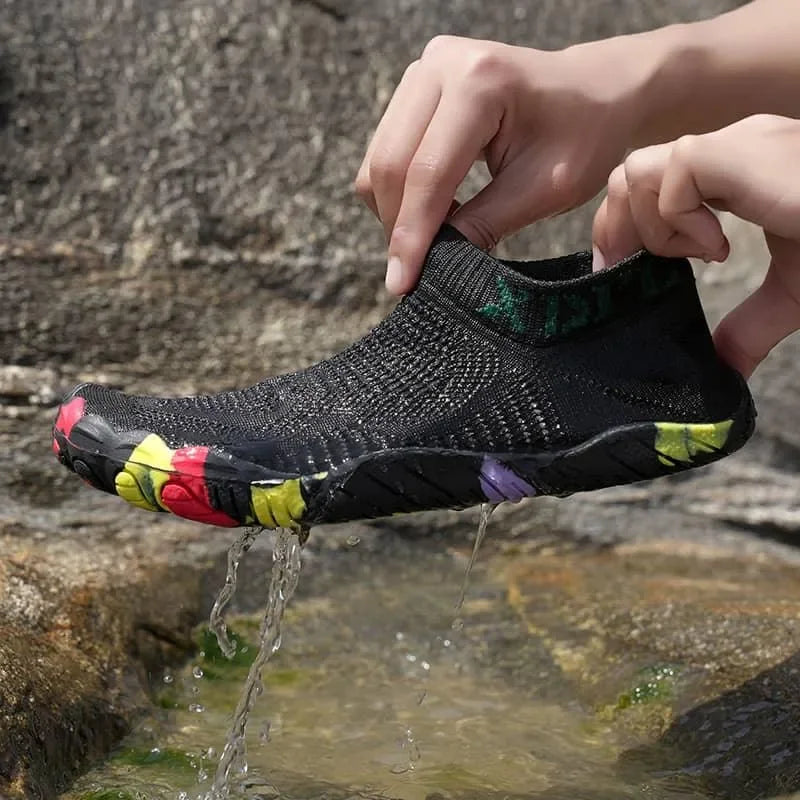 A black knit slip-on water shoe being held over flowing water outdoors, with colorful floral accents on the sole and hands visible.