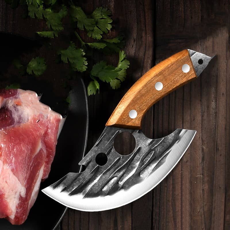 Curved meat cleaver with wooden handle on wooden table, raw meat and herbs nearby