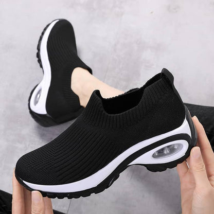 Black slip-on sneakers with white soles held by a hand on a gray background