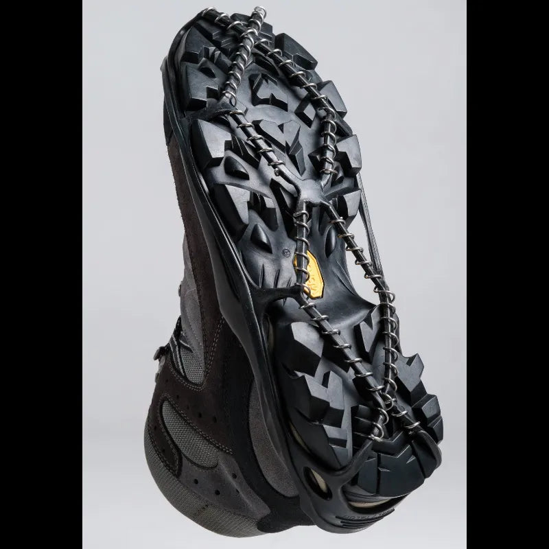 A black snowboard boot with a heavy-duty black metal crampon chain attached to the sole, shown against a light gray background.