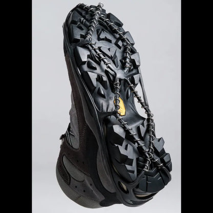 A black snowboard boot with a heavy-duty black metal crampon chain attached to the sole, shown against a light gray background.