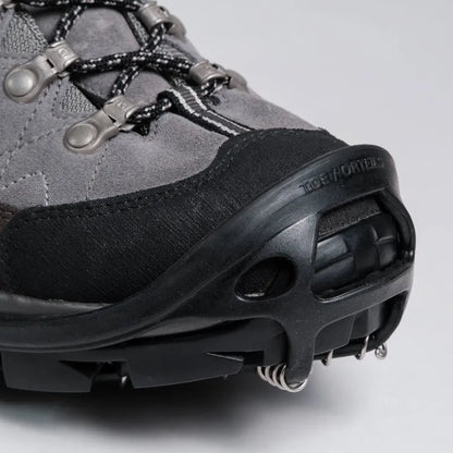 Close-up of a gray and black hiking boot with metal crampons attached to the sole, photographed on a light gray studio background. The brand name TORQOR is embossed on the black toe bumper.