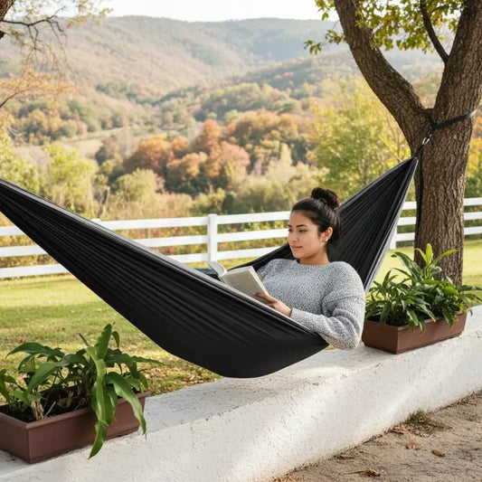 A woman relaxes in a black outdoor hammock strung between two trees beside a white fence, with a scenic autumn mountain landscape in the background.