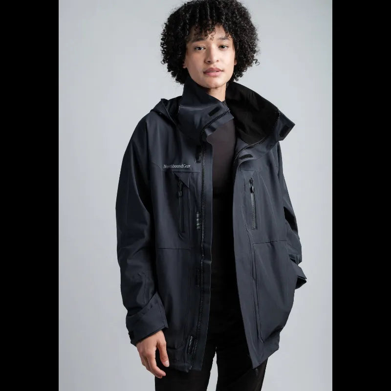 A model wearing a black hooded technical jacket with multiple zippered pockets, photographed against a neutral light gray studio background.