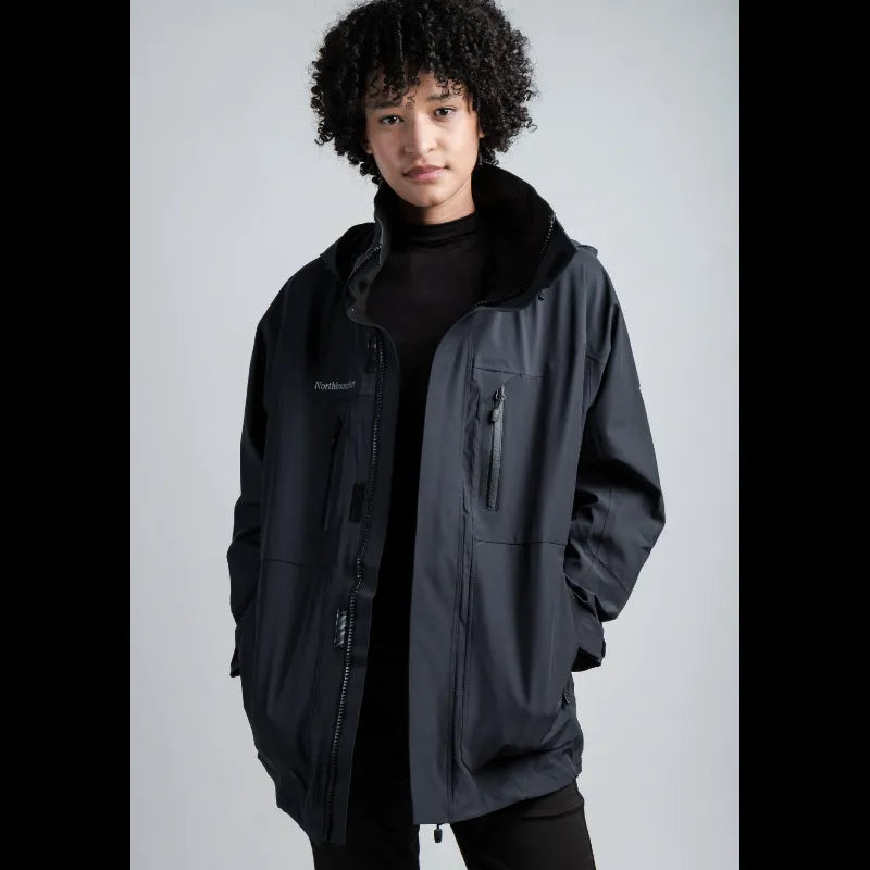 A model with curly black hair wearing a black hooded jacket with a black turtleneck underneath, set against a plain gray studio background.
