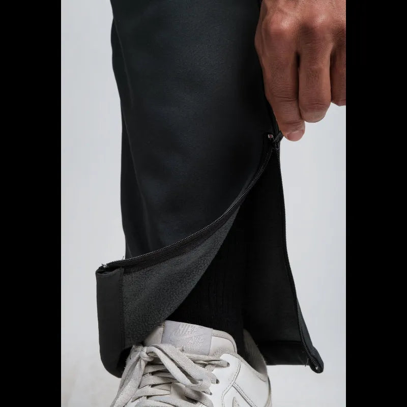 Close-up of a person wearing black pants, pulling a side zipper to reveal a fleece-lined pocket, with a white sneaker visible at the bottom against a light gray background.