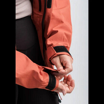 Close-up of a person wearing a coral orange technical jacket with black velcro cuffs, shown adjusting the sleeve. The jacket is paired with black pants against a light gray studio background.