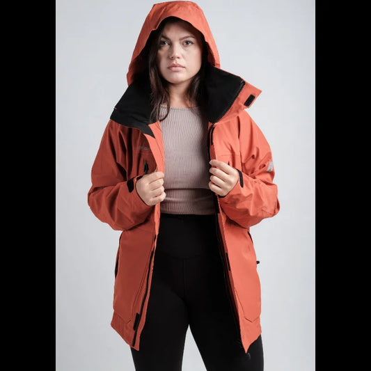 A woman wearing a hooded orange-red technical jacket with a black inner collar, styled over a ribbed mauve top and black leggings, set against a plain light gray studio background.