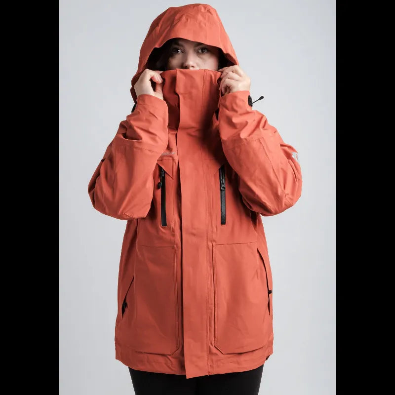 A person wearing a hooded orange jacket, holding the collar up with both hands against a light gray studio background.