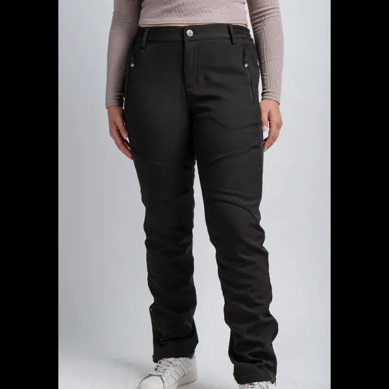 A model wearing black technical snow pants with zippered side pockets and reinforced knee panels, paired with a beige long-sleeve top and white sneakers, shown against a light gray studio background.