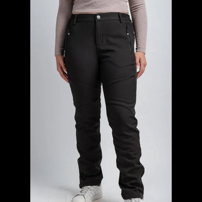 A model wearing black technical snow pants with zippered side pockets and reinforced knee panels, paired with a beige long-sleeve top and white sneakers, shown against a light gray studio background.