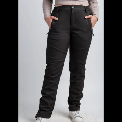A person wearing black technical pants with zippered pockets and a button waist, paired with a beige long-sleeve crop top and white sneakers, standing against a light gray studio background.