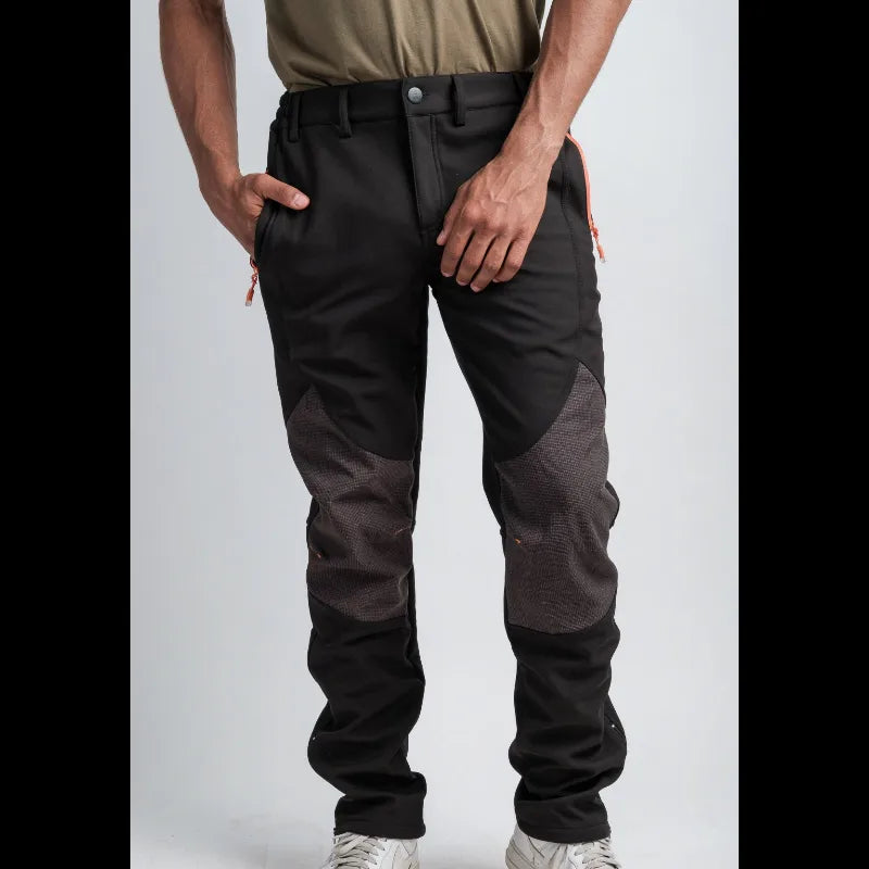 A person wearing black and dark gray color-blocked work pants with orange zipper pulls, paired with a tan shirt and white sneakers, shown from the waist down against a light gray studio background.