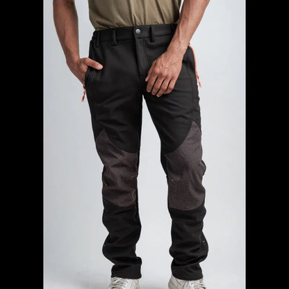 A person wearing black and dark gray color-blocked work pants with orange zipper pulls, paired with a tan shirt and white sneakers, shown from the waist down against a light gray studio background.