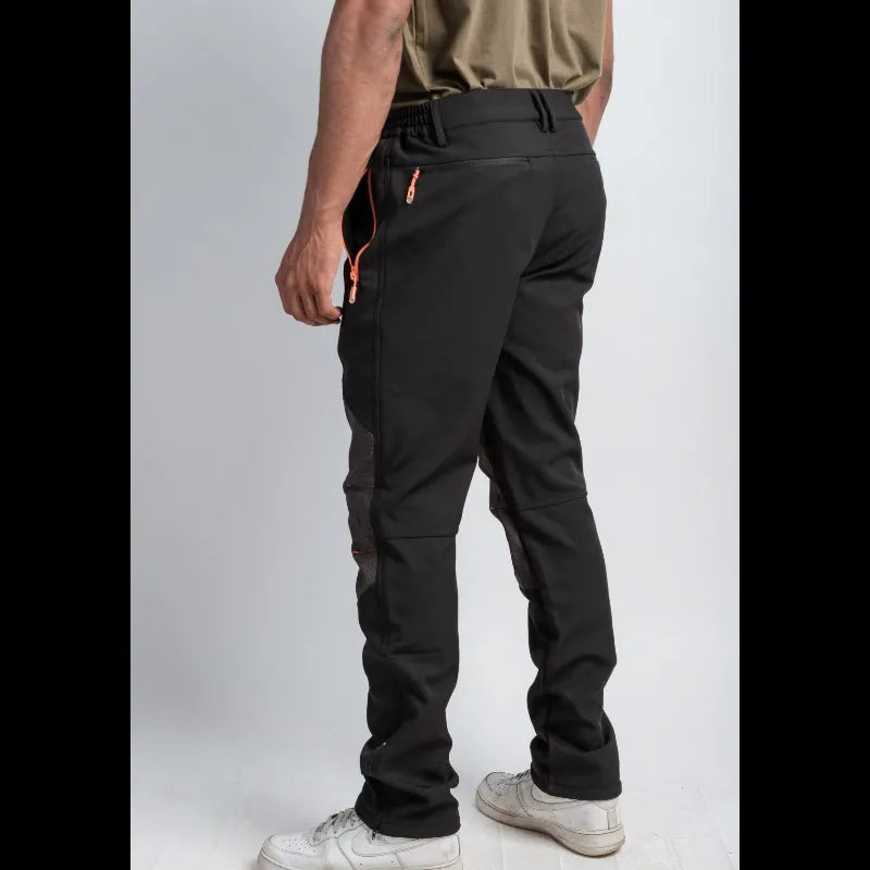 A person shown from the back wearing black technical pants with orange zipper accents, paired with a khaki shirt and white sneakers, against a light gray studio background.