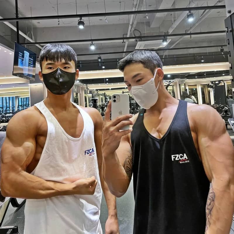 Two muscular individuals in a gym taking a selfie, wearing masks and tank tops with 'FZCA' branding.
