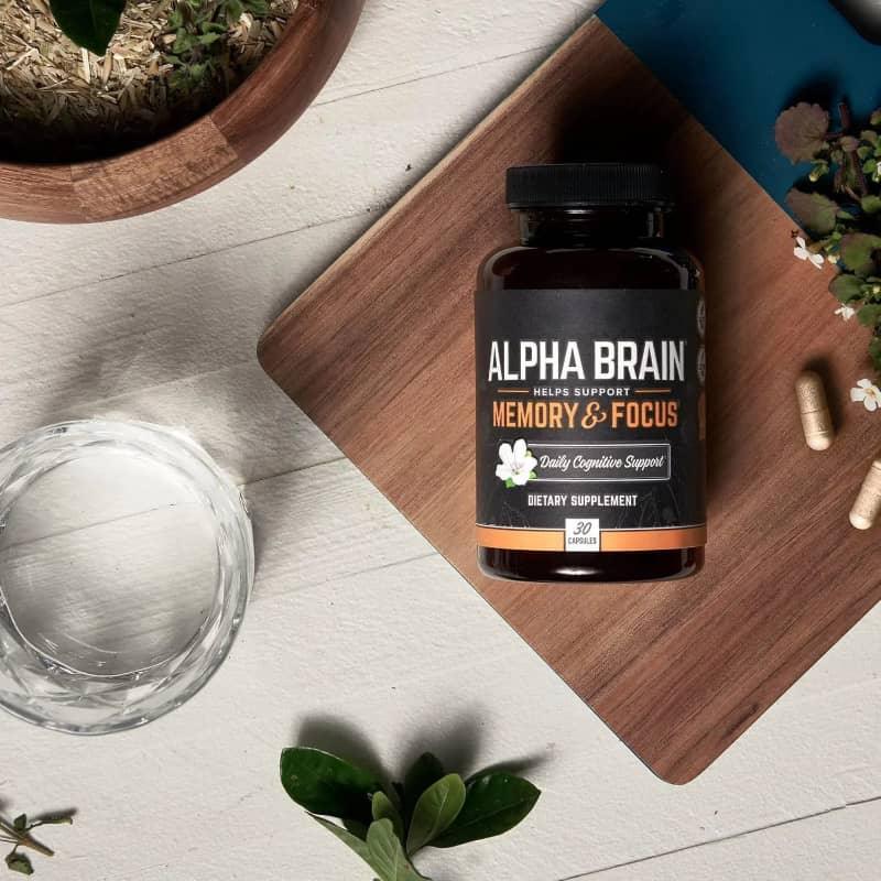 Alpha Brain memory and focus dietary supplement bottle with capsules, glass of water, and plants on a wooden board