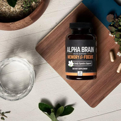 Alpha Brain memory and focus dietary supplement bottle with capsules, glass of water, and plants on a wooden board