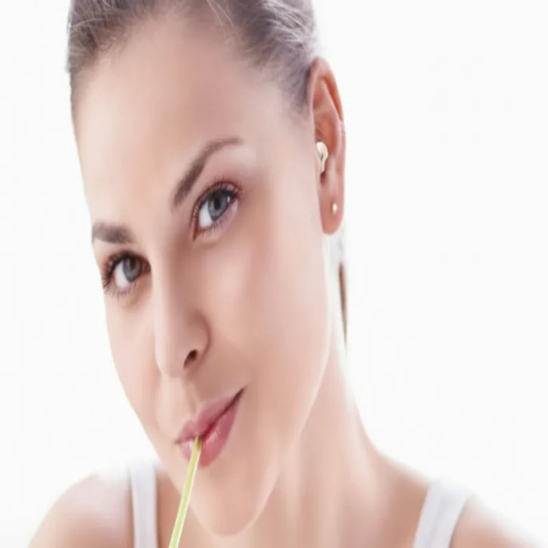 A close-up portrait of a woman with her hair pulled back, wearing small gold stud earrings and holding a yellow straw against her lips, set against a bright white background.