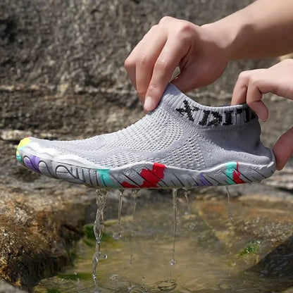 A gray knit slip-on water shoe being held over flowing water outdoors, with colorful accents on the sole and a visible brand mark on the collar.