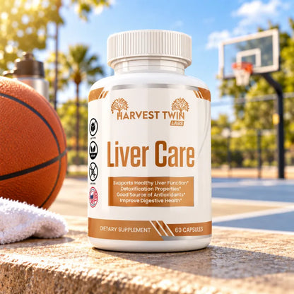 A white bottle of Harvest Twin Labs Liver Care dietary supplement stands on a stone surface outdoors at a basketball court, with a basketball, towel, and blurred court background visible.