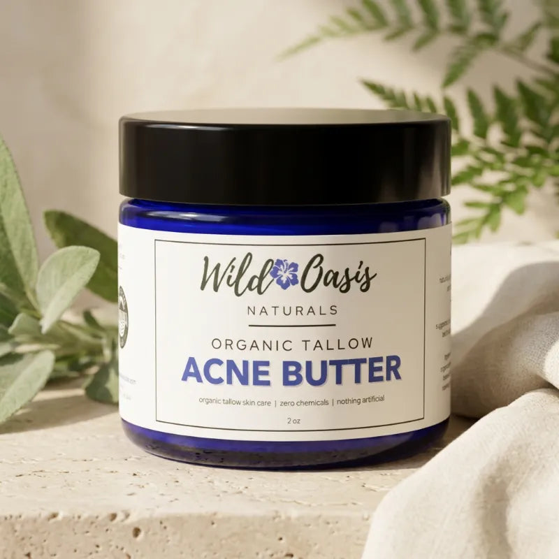 A jar of Wild Oasis Naturals Organic Tallow Acne Butter, featuring a dark blue glass jar, black lid, and white label, styled on a stone surface with soft green leaves and a beige cloth.