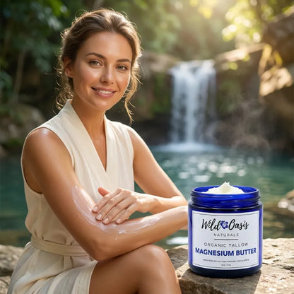 A woman in a cream robe sits outdoors by a waterfall, applying a white cream to her arm, with a jar of Wild Cow's Naturals Organic Tallow Magnesium Butter placed on the rock beside her.